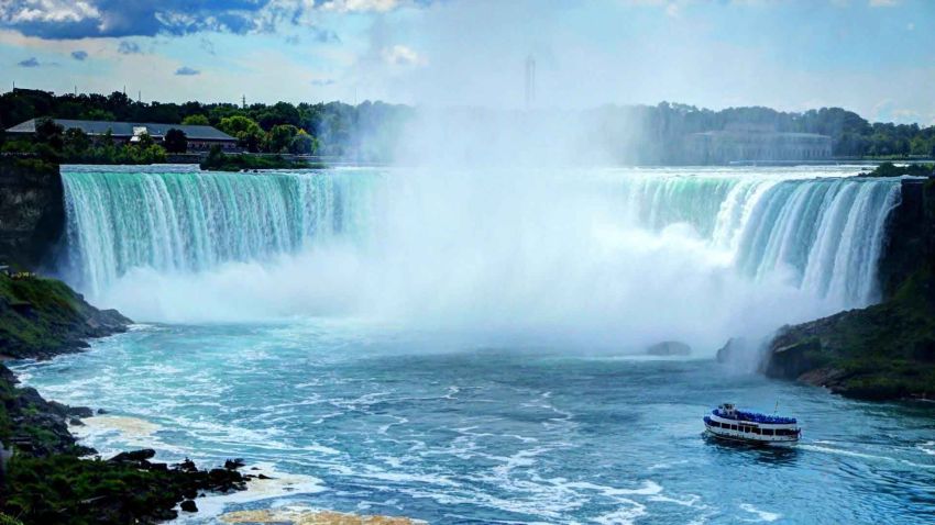 A powerful image of Niagara Falls, showcasing the massive Horseshoe Falls with a boat tour passing close to the base. The water is a vibrant turquoise, and mist rises dramatically from the falls.